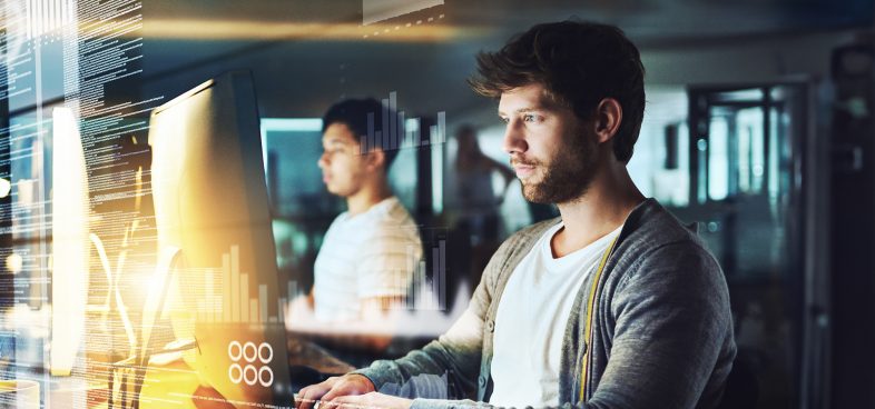 Two people working in a modern office environment with one person concentrating on a computer screen displaying code while another person works in the background.